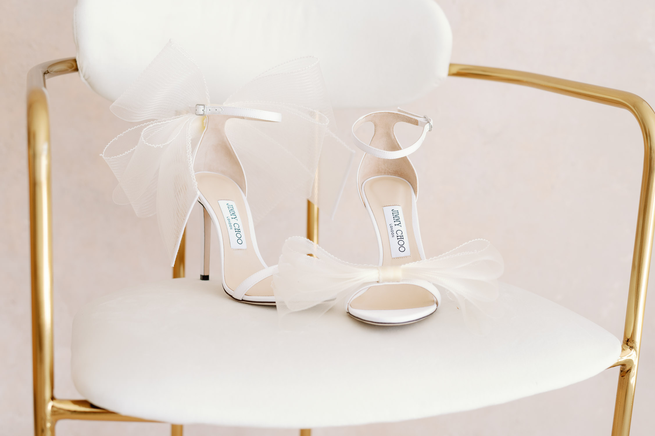 White Jimmy Choo shoes with large bows sit in a chair.