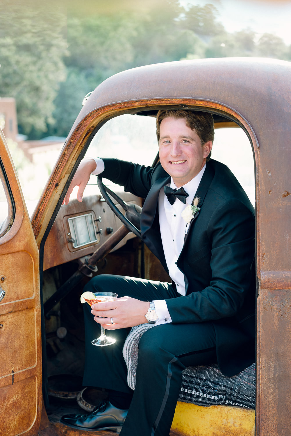 Groom leans out of the driver's side door of an antique truck while holding a cocktail.