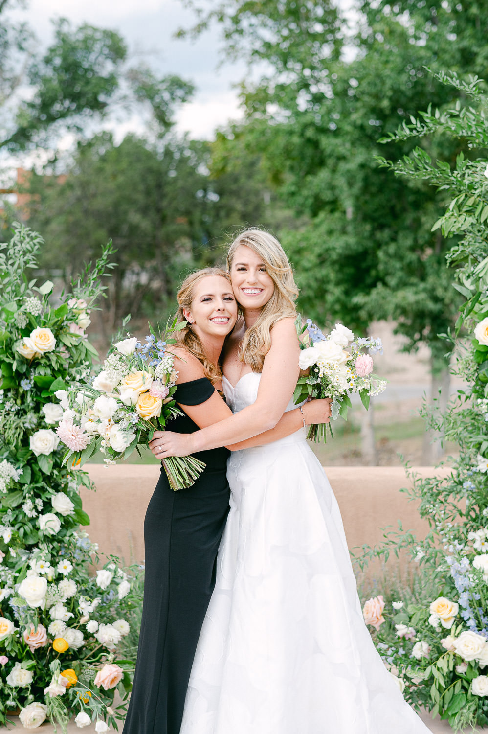 Bride and sister hug in front of a floral wedding arch.